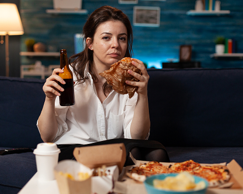 woman eating unhealthy burger and drinking alcohol on couch